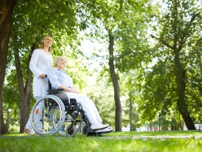 Woman in white pushing lady in wheelchair through the park.
